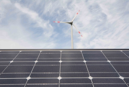 Windrad mit rot markierten Rotorblättern hinter einer großen Solaranlage unter bewölktem Himmel