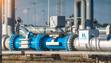 Blaue und weiße Rohrleitungen mit Ventilen und einem weißen Kasten mit der Aufschrift 'H2 HYDROGEN' in einer Industrieanlage unter blauem Himmel.