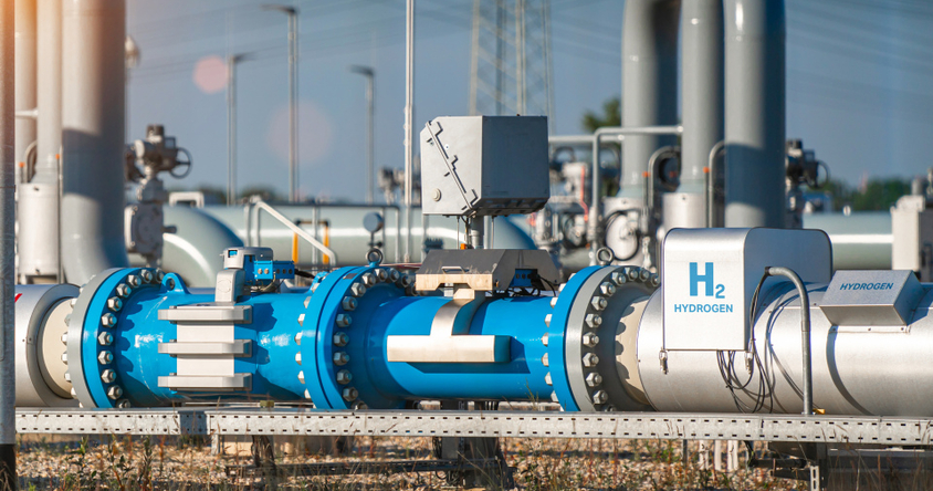 Blaue und weiße Rohrleitungen mit Ventilen und einem weißen Kasten mit der Aufschrift 'H2 HYDROGEN' in einer Industrieanlage unter blauem Himmel.